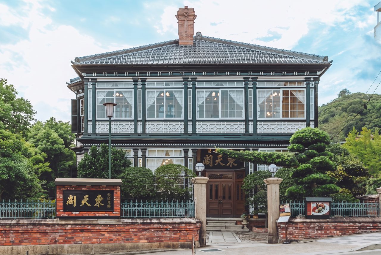 神戸・北野に建つ東天閣の洋館。緑色の木造二階建てで、煉瓦の塀と門柱、松の植栽が手前にある